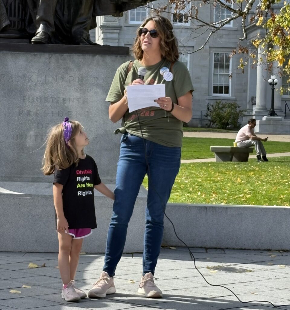 Julie speaks into a microphone at ABLE NH's 2024 Disability Justice Parade, as her young daughter stands beside her and watches her.