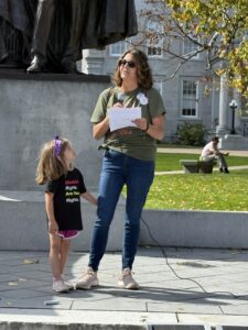 Photo of Julie speaking with a microphone as her young daughter stands beside her, watching