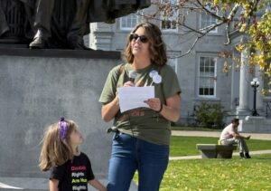 Photo of Julie speaking with a microphone as her young daughter stands beside her, watching