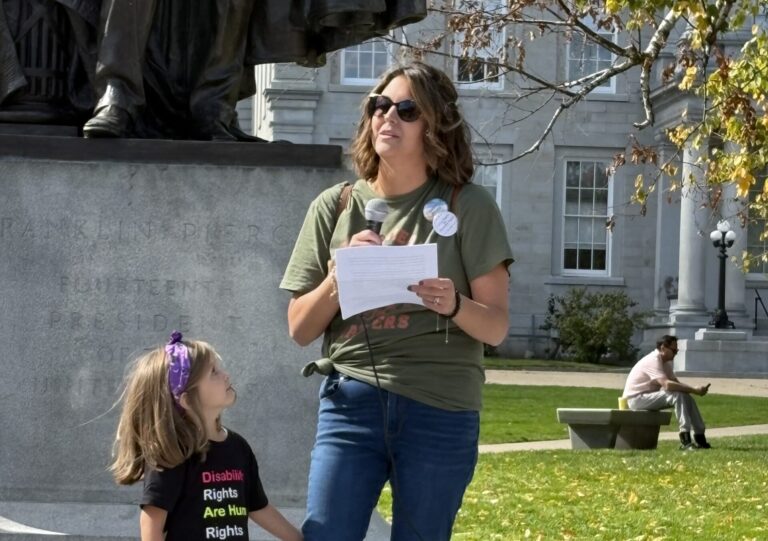 Photo of Julie speaking with a microphone as her young daughter stands beside her, watching