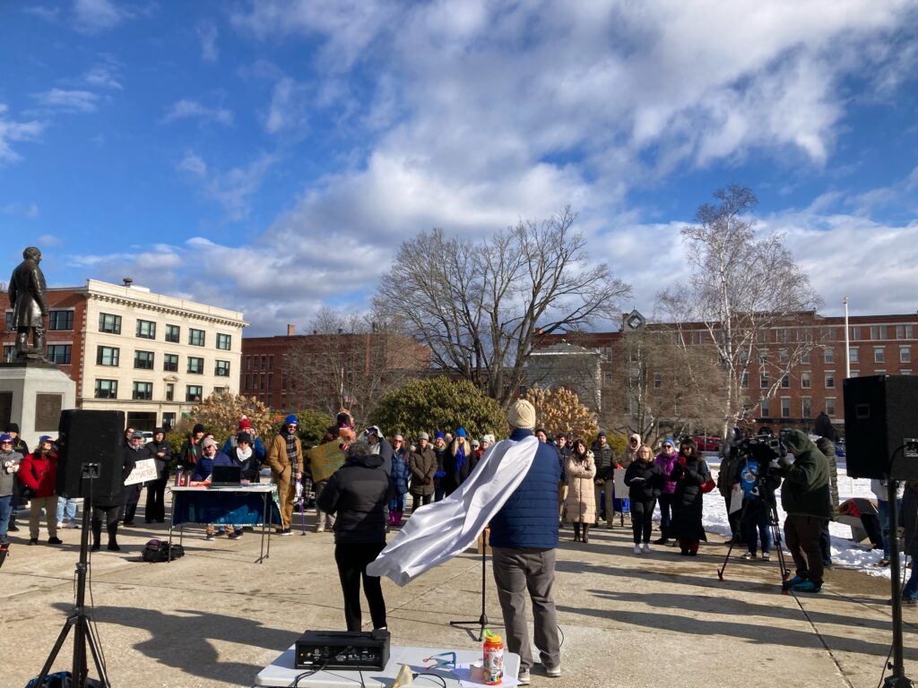 Executive Director Louis wearing a blue coat and winter hat wearing a silver cape speaking in front of an audience